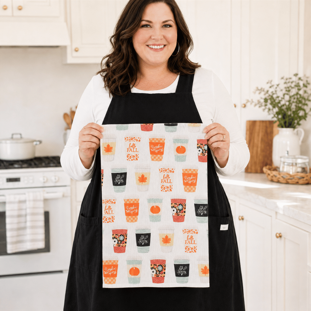 A woman wearing a Plus Size black apron holds up a white kitchen towel featuring a colorful 'Fall is here' pattern with mugs, pumpkins, and autumn motifs, set against a bright, modern kitchen background.