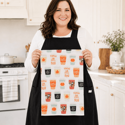 A woman wearing a Plus Size black apron holds up a white kitchen towel featuring a colorful 'Fall is here' pattern with mugs, pumpkins, and autumn motifs, set against a bright, modern kitchen background.